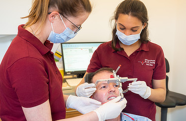Ein Patient bekommt von einer Zahnärztin und einer Zahnarzthelferin das Gesicht mit einem Gesichtsbogen vermessen.