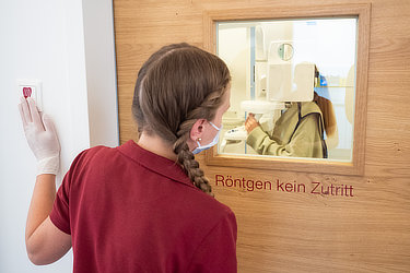 Eine Frau blickt durch das Fenster einer Tür in den Röntgenraum, wo eine Patientin gerade den Kiefer geröntgt bekommt.